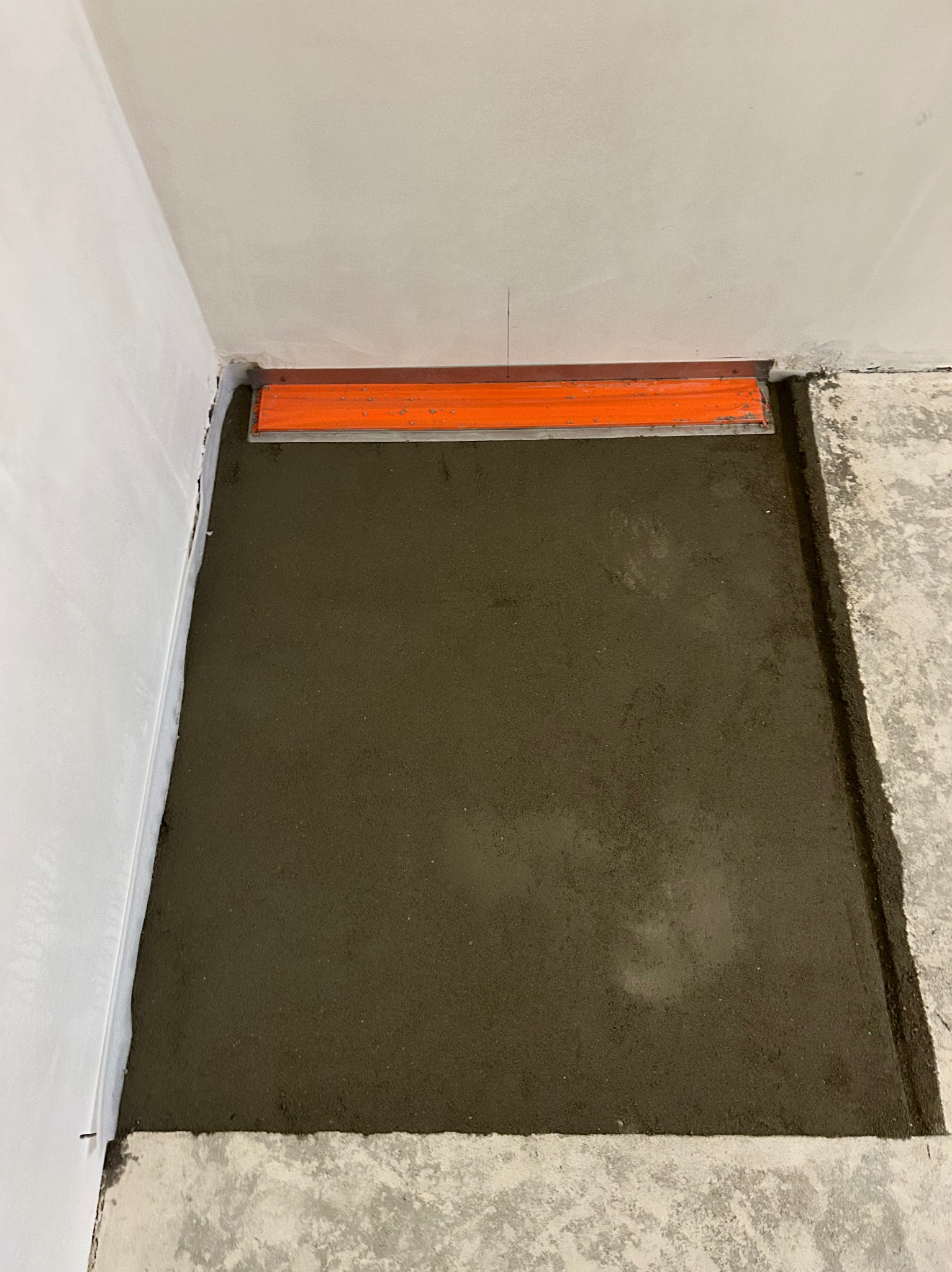 Wyrównywanie wylewki podłogowej w rogu pomieszczenia za pomocą pomarańczowej łaty. Widoczny fragment ściany i surowa podłoga.