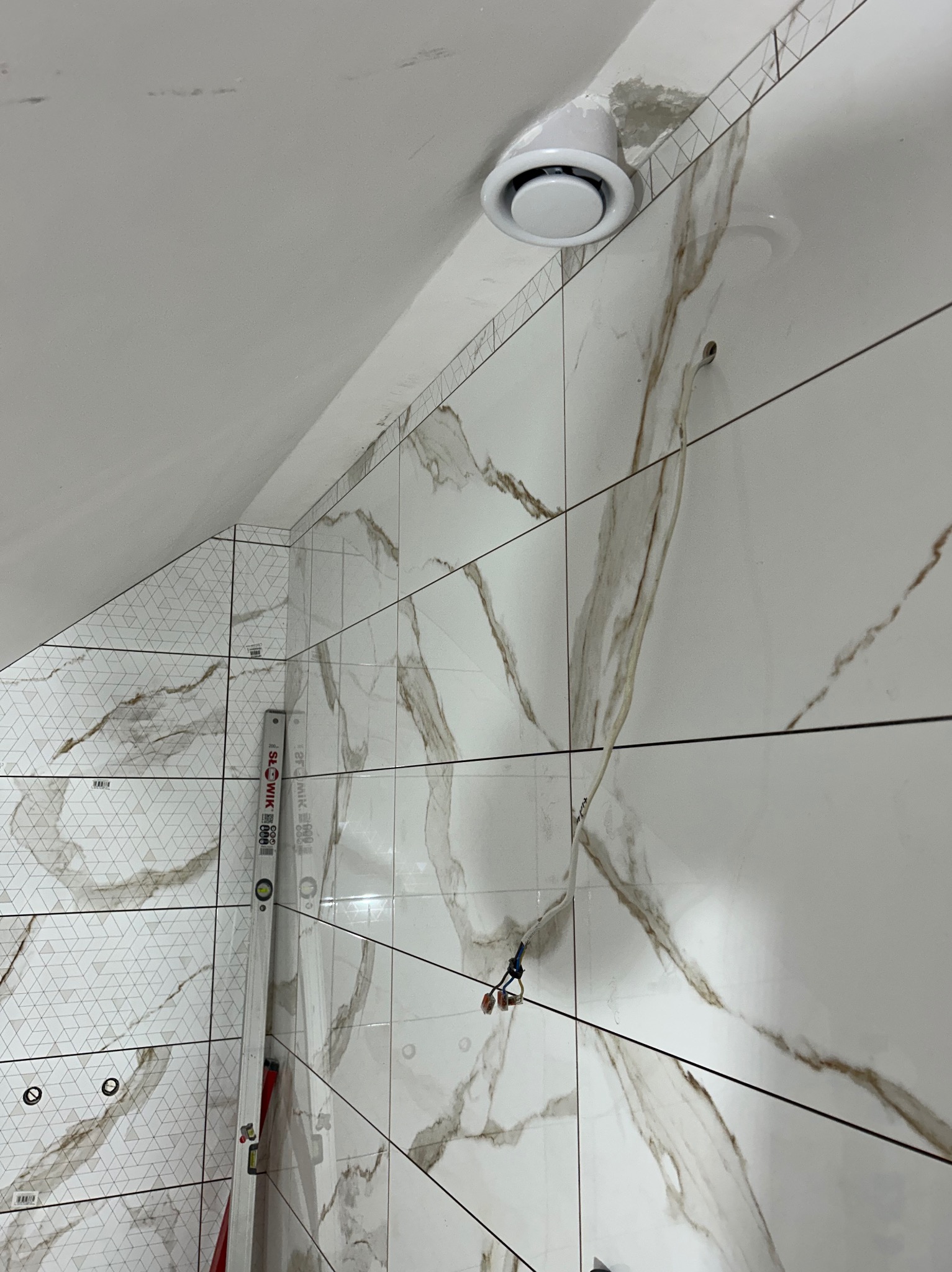 Białe płytki imitujące marmur na ścianie łazienki, widoczny wentylator sufitowy, poziomica i wystające przewody elektryczne. Ukośny sufit.