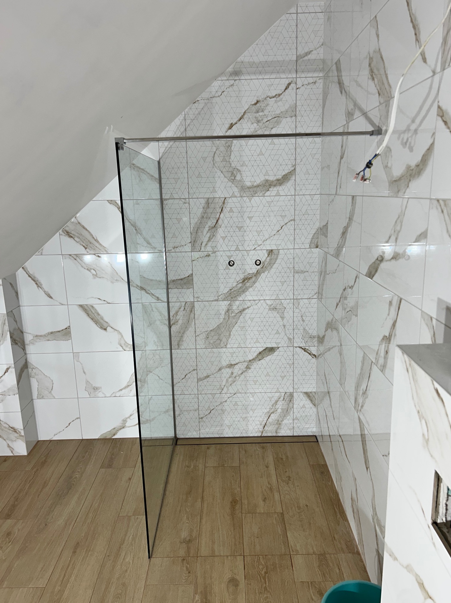 Ukończona łazienka na poddaszu: marmurowe płytki ścienne z geometrycznym wzorem, szklana kabina prysznicowa, drewnopodobna podłoga. Widoczne wystające kable.