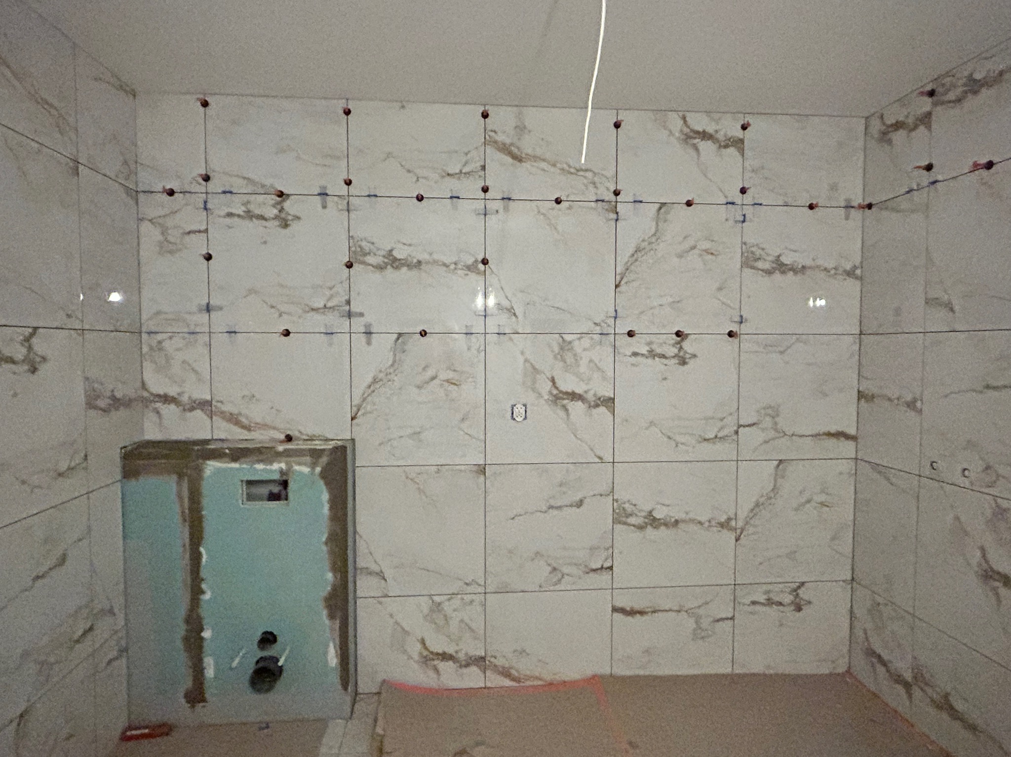 Ściana w łazience z ułożonymi płytkami imitującymi marmur, z widocznymi krzyżykami dystansowymi i elementami instalacji podtynkowych. Etap prac wykończeniowych.