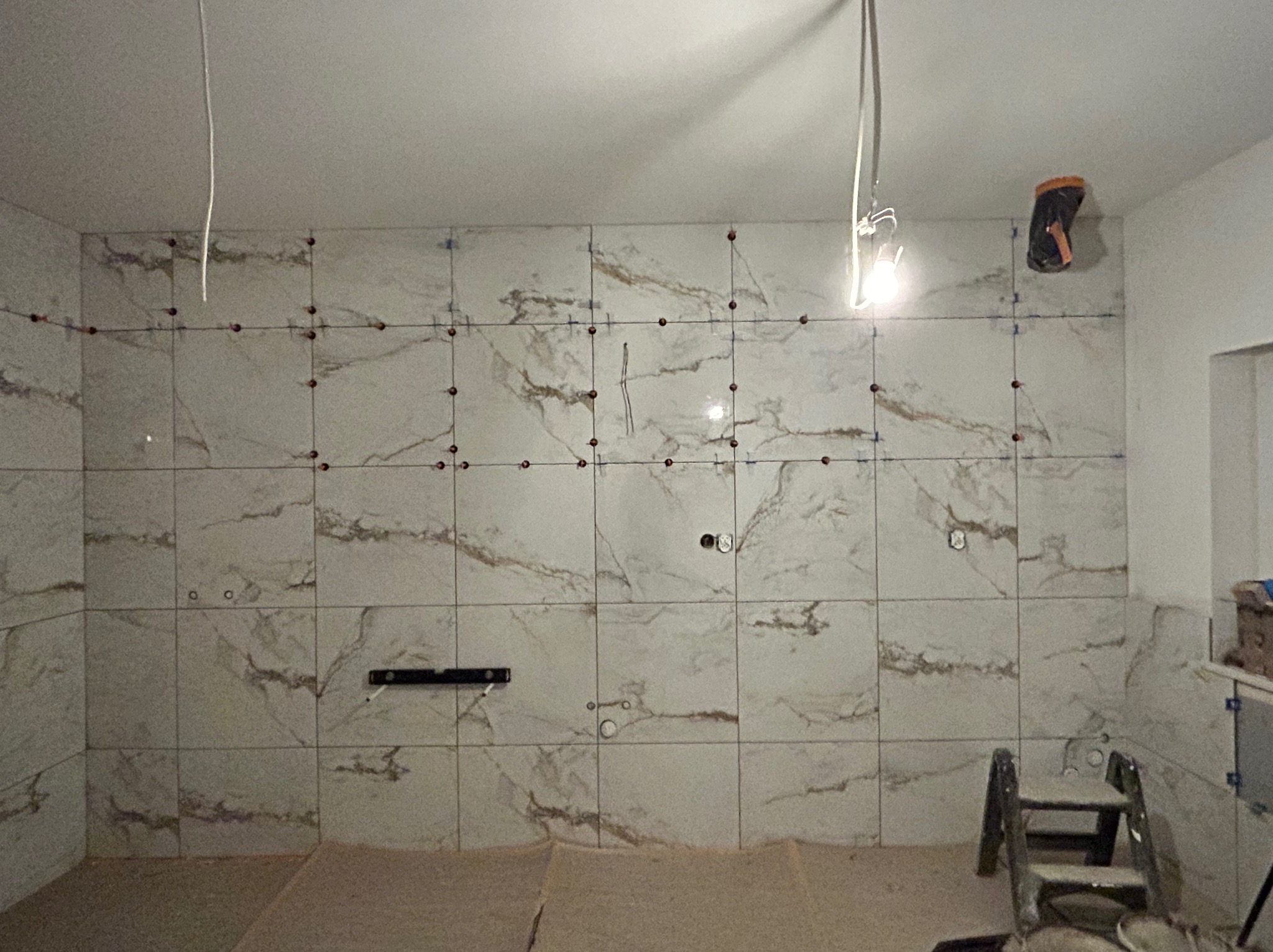 Ściana w trakcie układania płytek imitujących marmur, z widocznymi krzyżykami dystansowymi i poziomicą. W tle drabina i narzędzia glazurnicze.