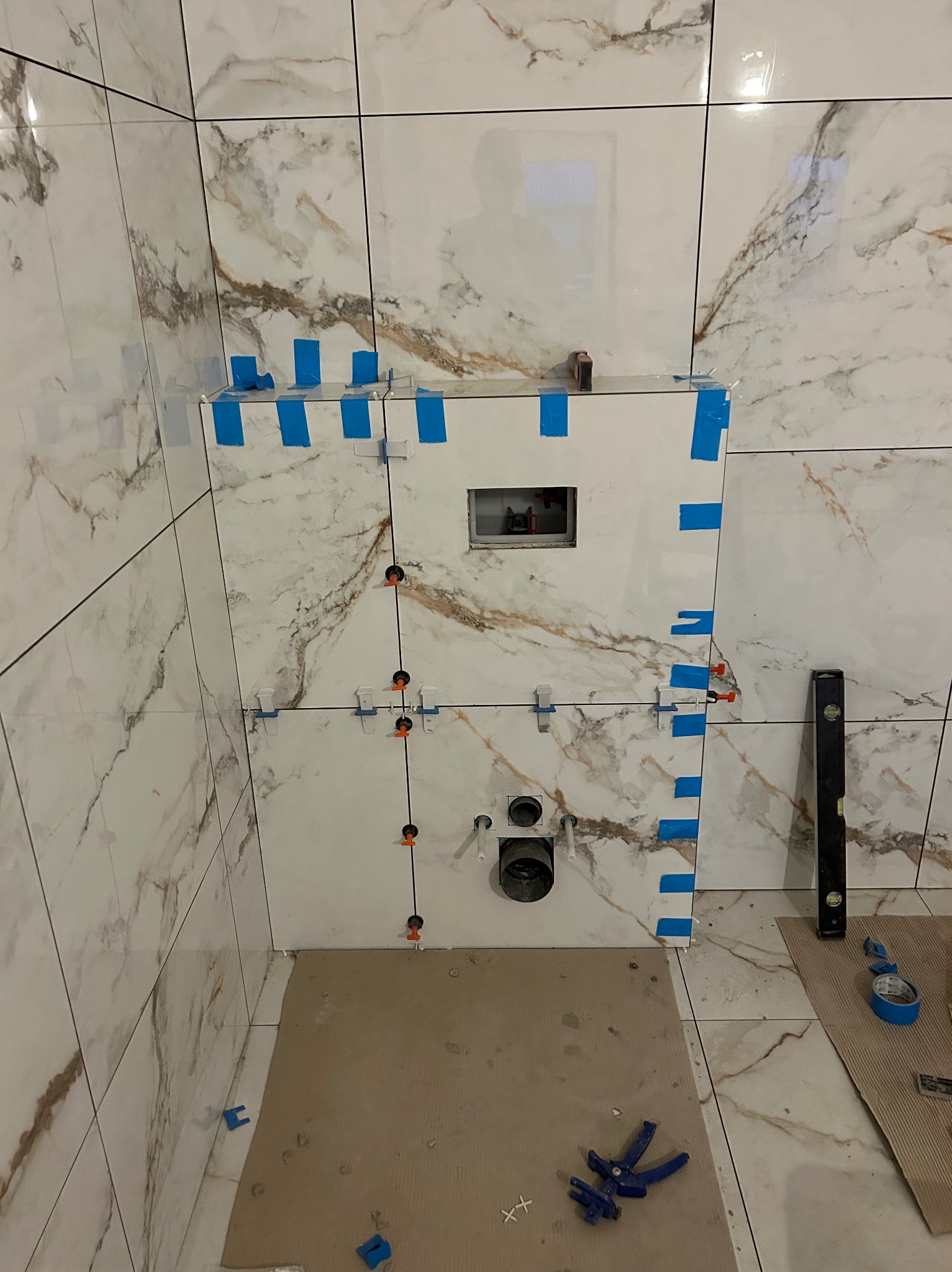 Montaż płytek imitujących marmur w łazience, widoczne poziomica, taśma malarska i narzędzia glazurnicze. Etap prac z zamontowaną instalacją podtynkową WC.