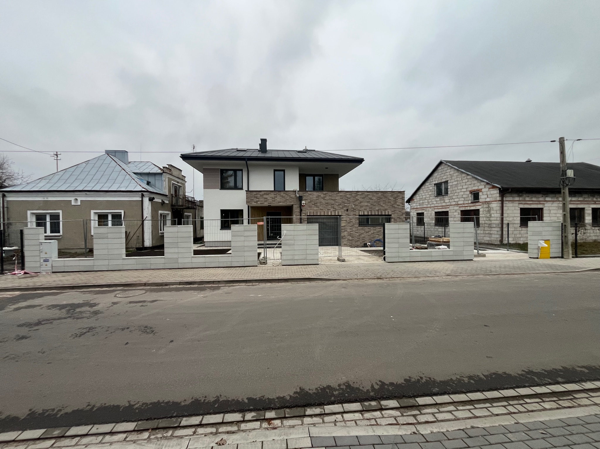 Wznoszenie ogrodzenia z prefabrykatów betonowych przed trzema różnymi domami w zabudowie szeregowej, widok z ulicy. Etap budowy, widoczne elementy konstrukcyjne.