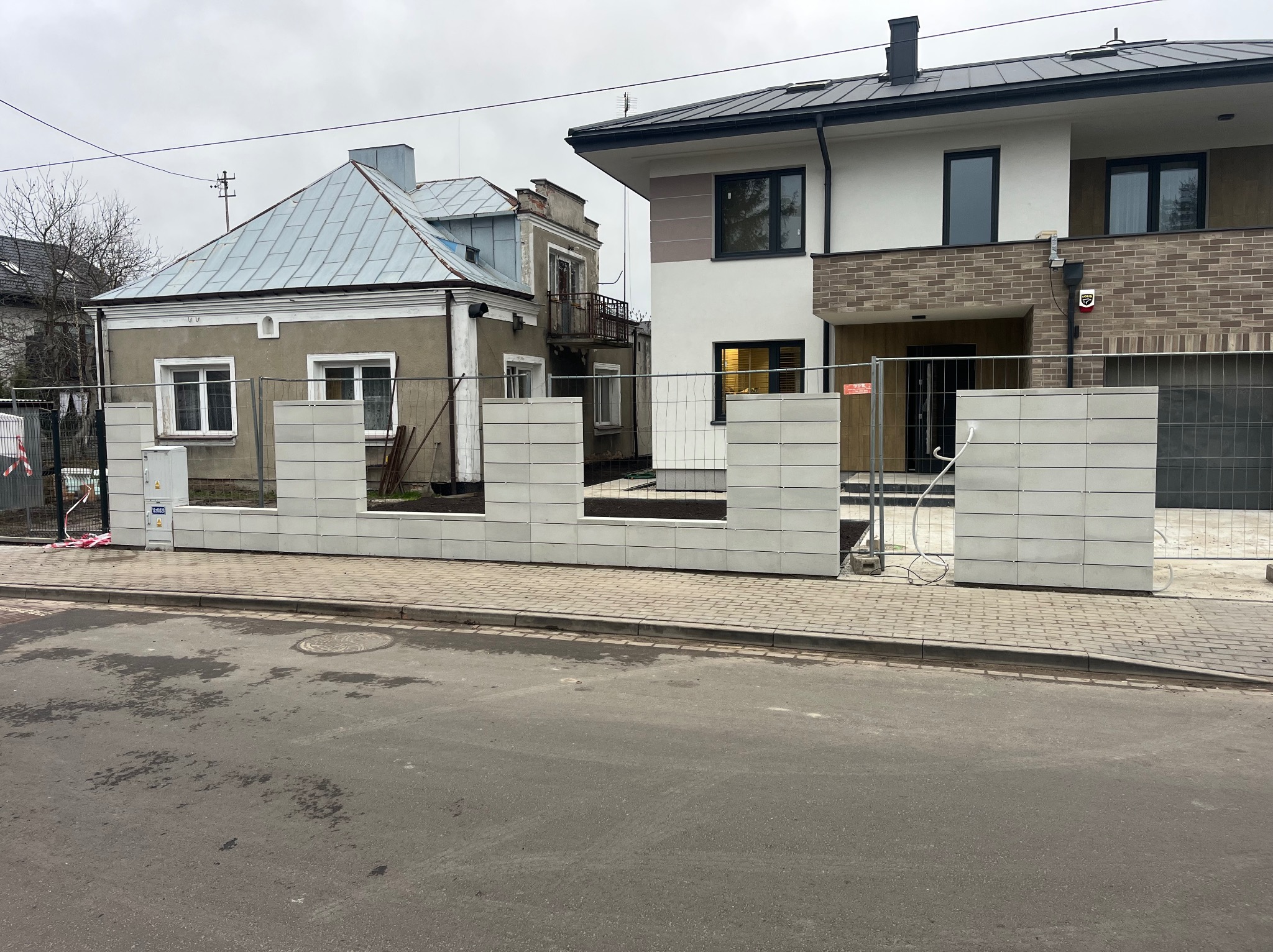 Wznoszony, nowoczesny, modułowy mur z jasnych betonowych bloczków, częściowo otoczony tymczasowym ogrodzeniem budowlanym, na tle dwóch budynków.