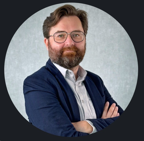 Portret mężczyzny z brodą w okularach, ubrany w marynarkę i koszulę, z założonymi rękami na tle jasnej ściany. Ujęcie w okręgu.