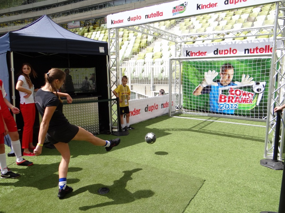 Kobieta kopie piłkę na tle stoiska z bramką i grafiką bramkarza, w tle trybuny stadionu. Event firmowy z atrakcjami sportowymi i namiotem.