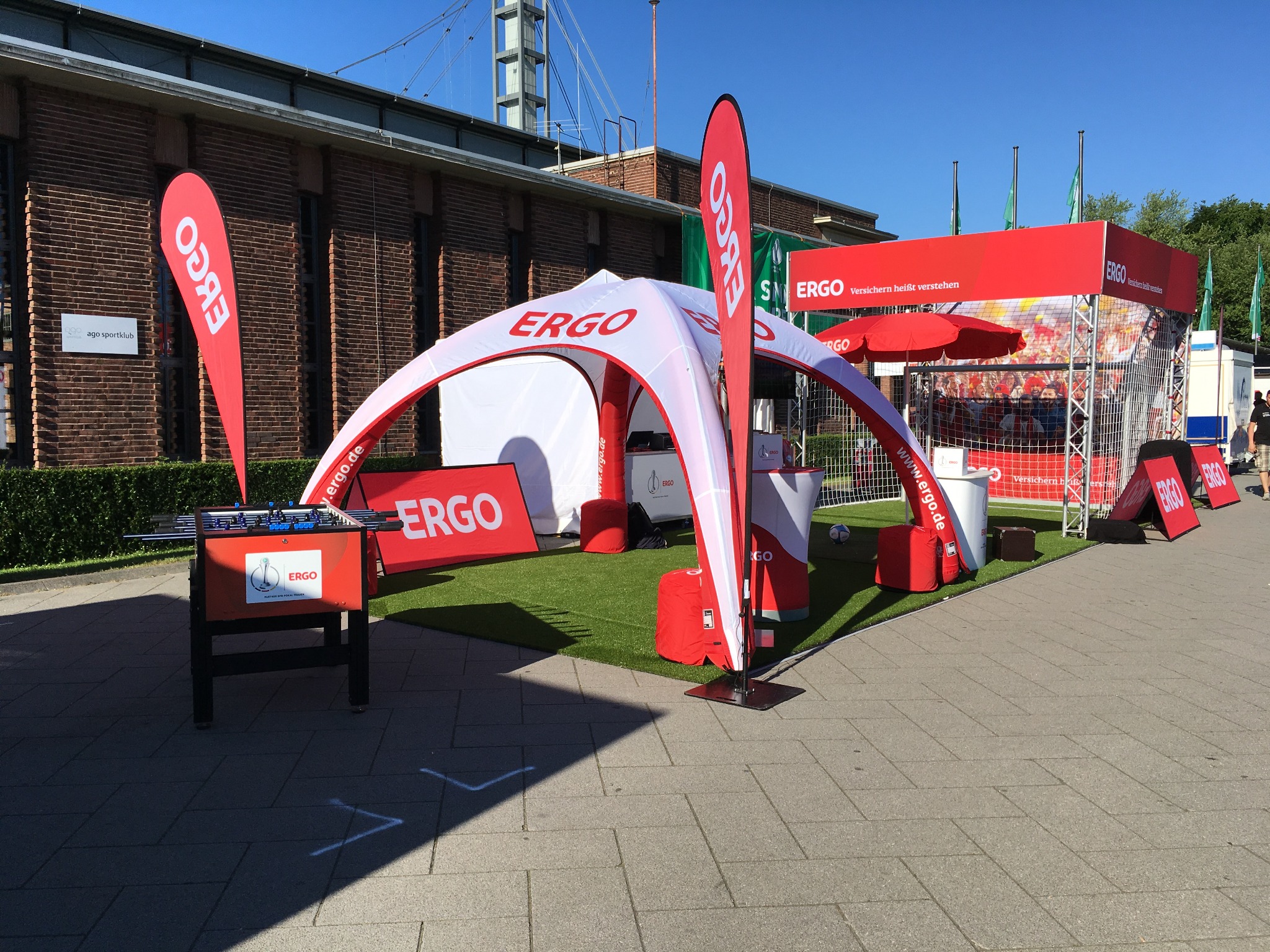 Stoisko promocyjne ERGO z namiotem, piłkarzykami i piłką nożną na sztucznej trawie. Czerwono-biała kolorystyka, logo ERGO widoczne na elementach stoiska. Budynek w tle.
