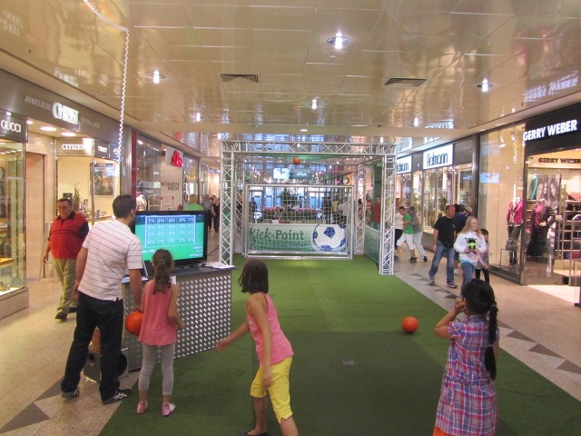 Interaktywna strefa rozrywki 'Kick Point' w centrum handlowym: dzieci grają w piłkę nożną, obok ekran z wynikami, w tle widoczni przechodnie i sklepy.