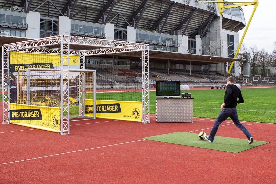 Interaktywna strefa kibica z bramką, ekranem i konstrukcją z logo BVB na stadionie. Mężczyzna przygotowuje się do strzału na tle trybun.