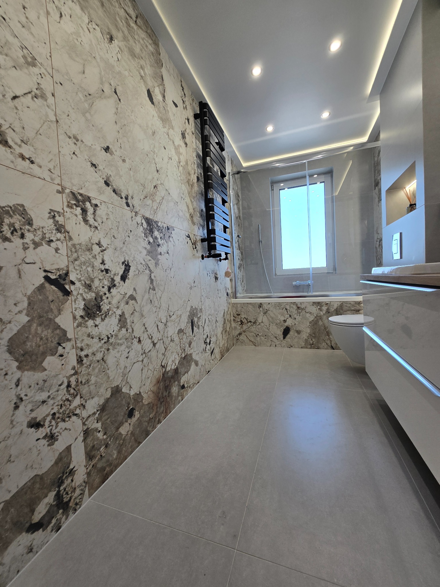 Elegancka łazienka z marmurowymi płytkami na ścianach, szklaną kabiną prysznicową i grzejnikiem drabinkowym. Nowoczesne oświetlenie sufitowe.