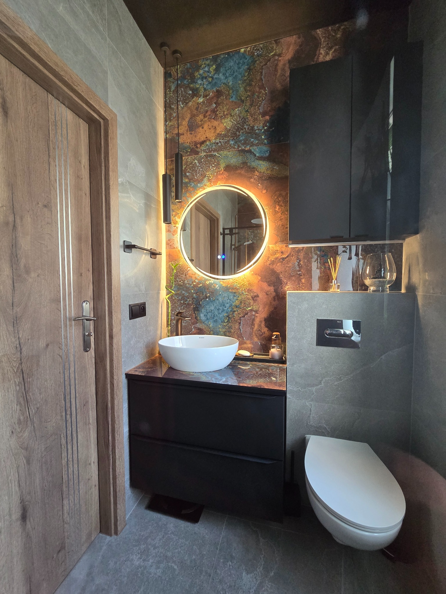 Nowoczesna łazienka z umywalką nablatową, okrągłym lustrem z podświetleniem LED i płytkami imitującymi kamień. Widoczna drewniana stolarka drzwiowa.