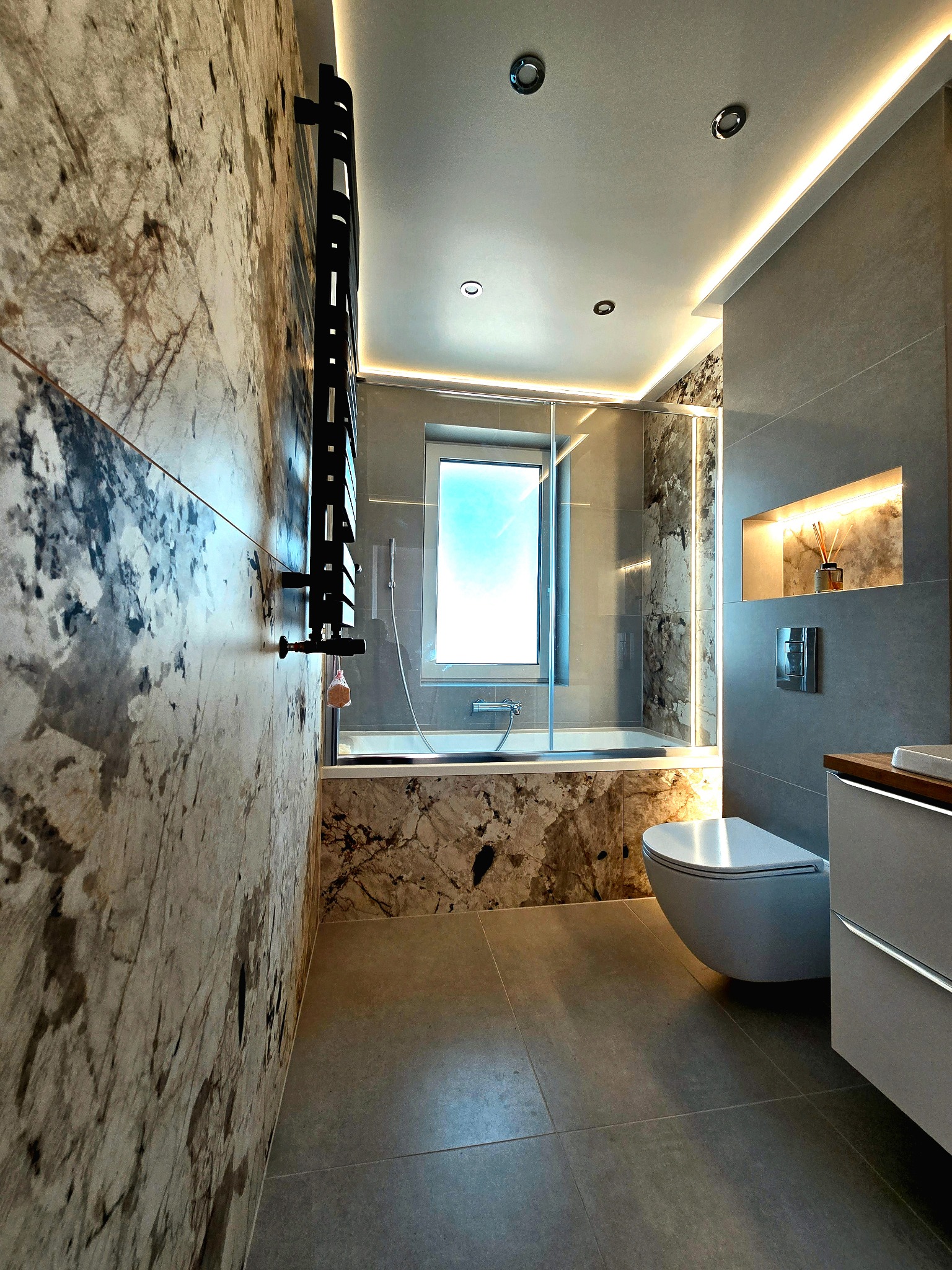 Elegancka łazienka z marmurowymi akcentami, podświetleniem LED i nowoczesną armaturą. Widok na wannę za szklaną ścianką i minimalistyczną toaletę.