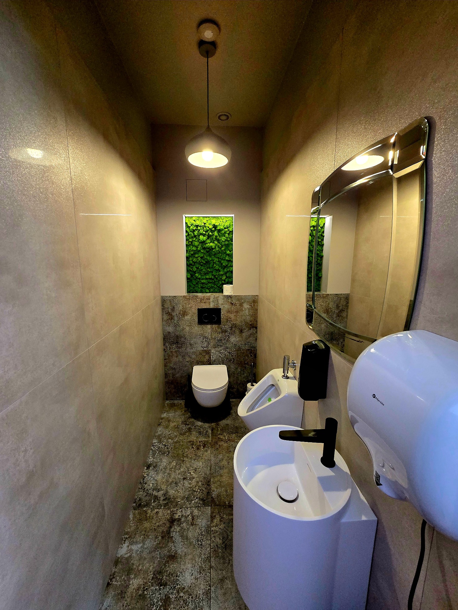 Nowoczesna toaleta publiczna z umywalką, pisuarami i toaletą. Ściany wyłożone płytkami w odcieniach beżu, podłoga w szarościach. W tle okno z zieloną roślinnością.