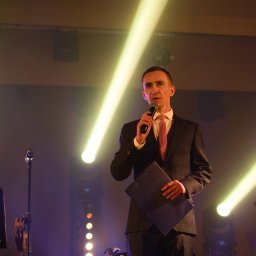 Dominik Rożek - Konferansjer w czarnym garniturze, z mikrofonem i teczką na tle sceny z reflektorami i choinką. Prezentacja wydarzenia.