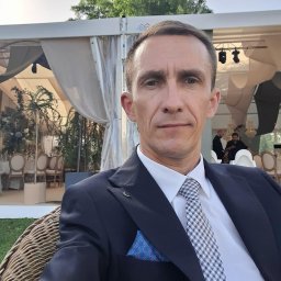 Dominik Rożek - Mężczyzna w garniturze siedzi na tle udekorowanego namiotu eventowego z krzesłami i roślinami, sugerując profesjonalną obsługę imprez.