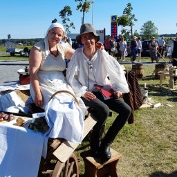 Dominik Rożek - Uśmiechnięta para w strojach historycznych siedzi na wozie z jedzeniem podczas plenerowej imprezy. W tle widoczni ludzie i budynek stacji benzynowej.