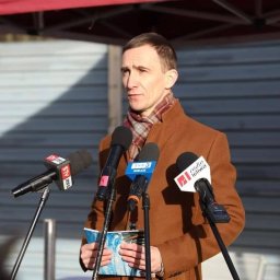 Dominik Rożek - Mężczyzna w brązowym płaszczu przemawia do mikrofonów z logotypami stacji radiowych i telewizyjnych, trzymając notatki. Ujęcie z zewnątrz, w dziennym świetle.