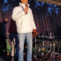 Dominik Rożek - Mężczyzna w białej kurtce i jeansach stoi na scenie z mikrofonem, w tle perkusja i sylwetki ludzi. Scena oświetlona, w tle drzewa.