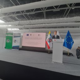 Dominik Rożek - Konferencja podsumowująca PROW 2014-2020 w Targach Kielce. Mężczyzna w garniturze przemawia zza mównicy, obok flagi UE i Polski. Banery informacyjne w tle.