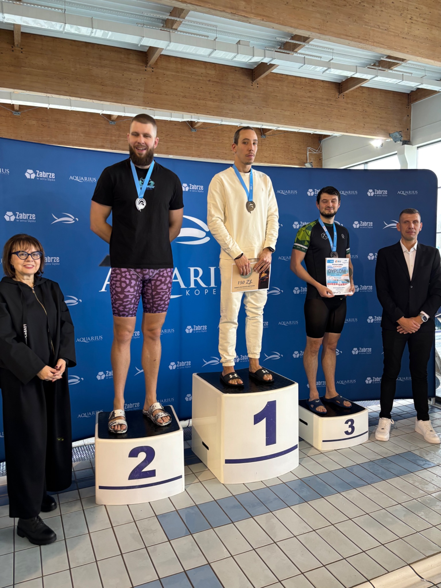 Zawodnicy z medalami na podium po zawodach pływackich w Aquarius Zabrze. Osoby na stopniach 1, 2 i 3, trenerzy obok. Dyplom i uśmiechy.