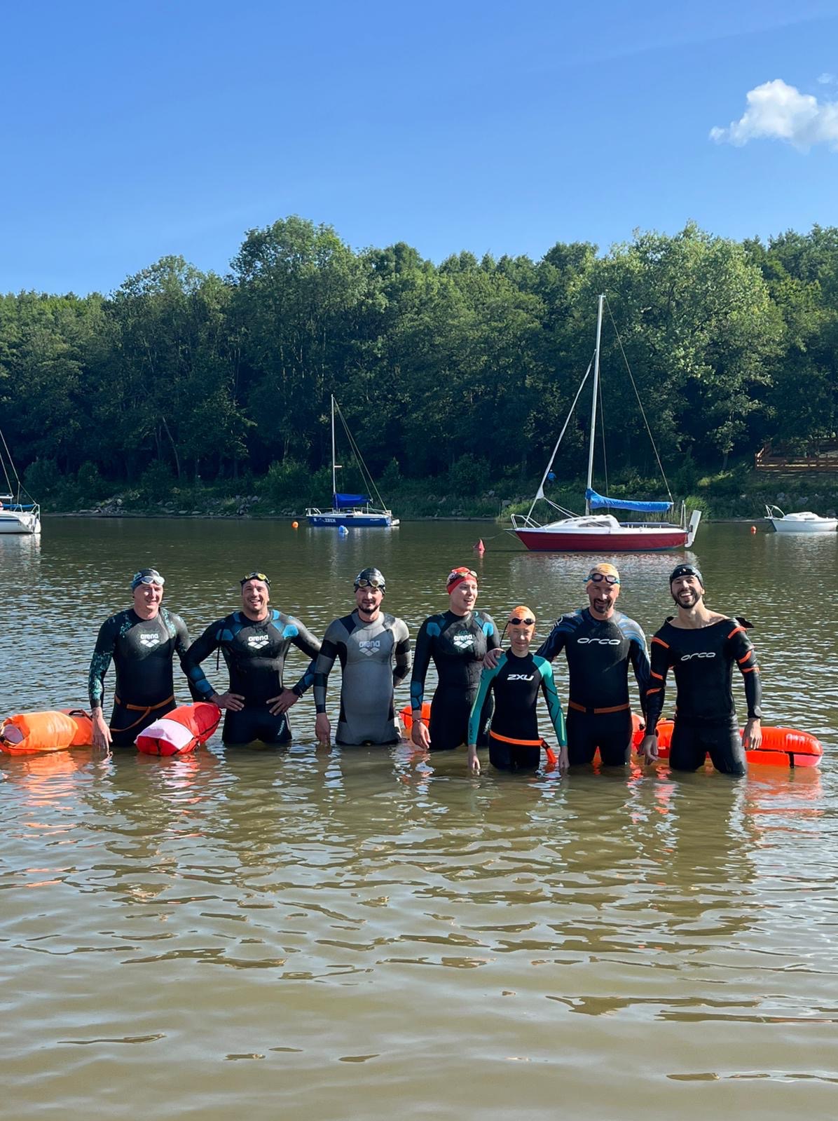 Grupa siedmiu osób w piankach stoi w wodzie jeziora, z bojkami asekuracyjnymi. W tle las i żaglówki pod błękitnym niebem. Uczestnicy zajęć z pływania na otwartych wodach.