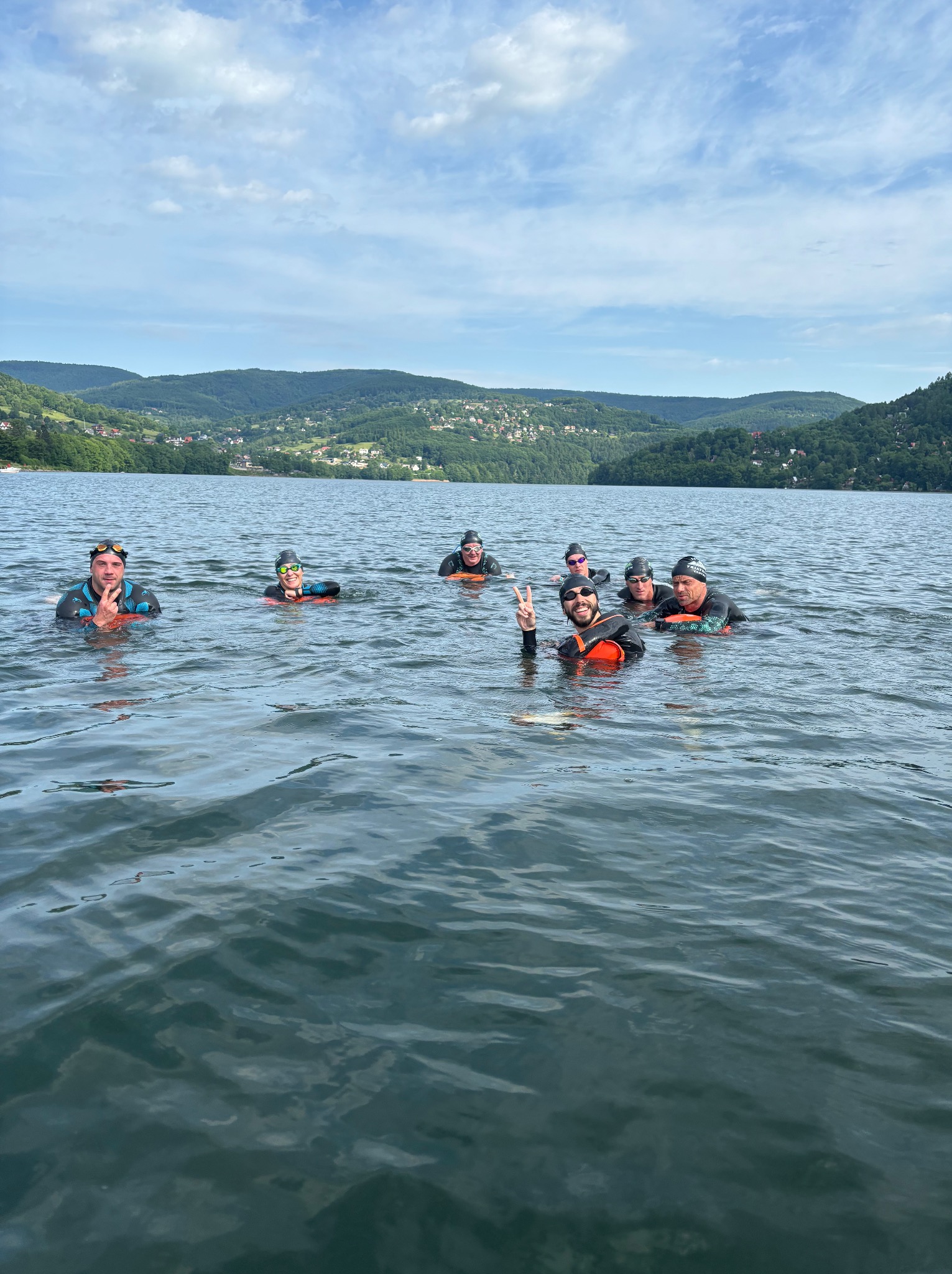 Grupa osób w piankach i czepkach pływa w jeziorze na tle zalesionych wzgórz i błękitnego nieba z chmurami. Uczestnicy kursu pozdrawiają, unosząc dłonie.
