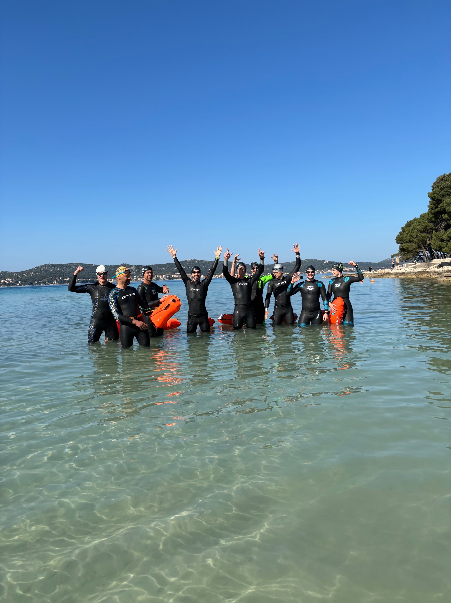 Grupa ośmiu pływaków w piankach, stojących w płytkiej, przejrzystej wodzie z pomarańczowymi bojkami, świętujących pod błękitnym niebem. Widok na wybrzeże w tle.