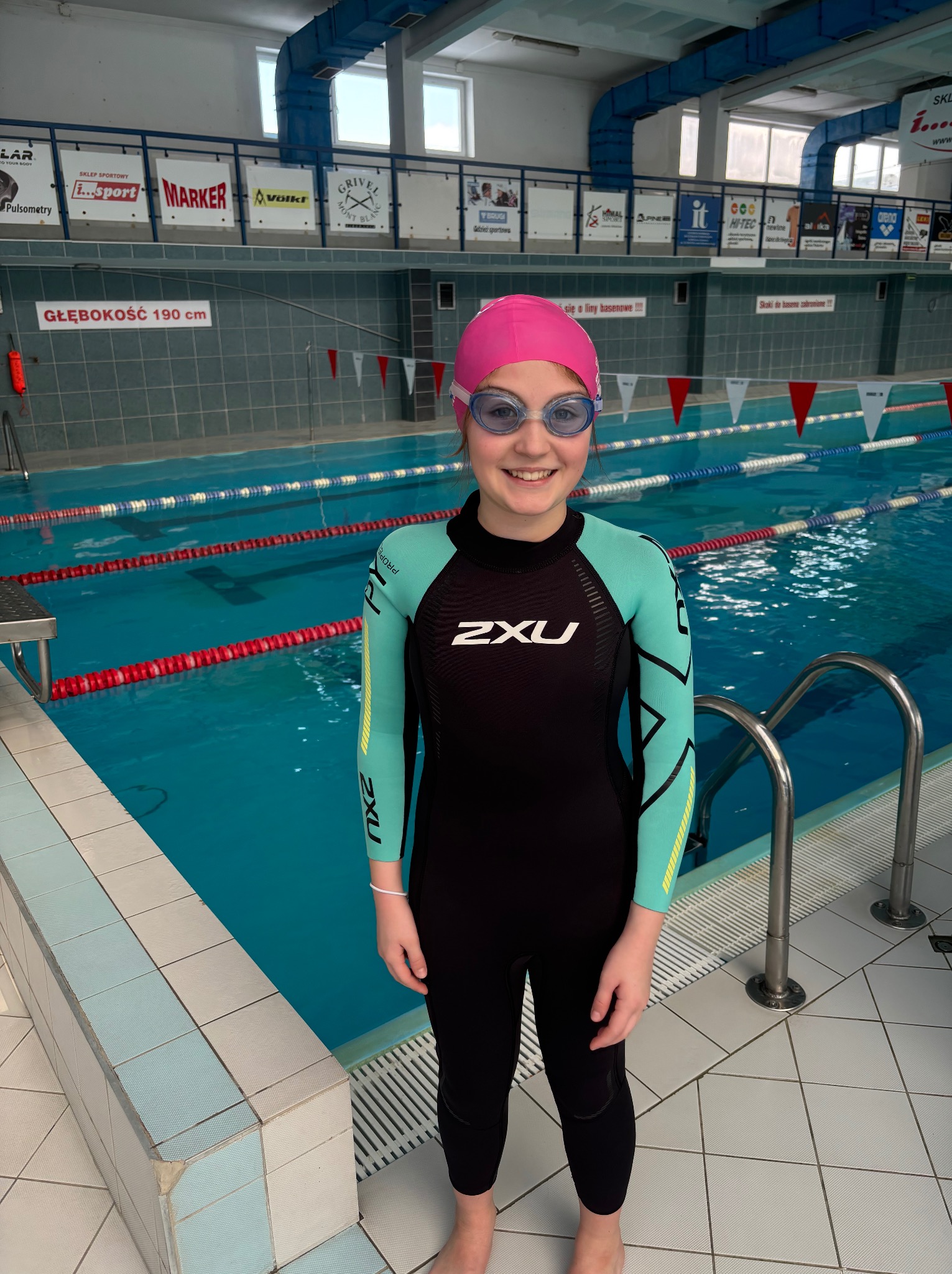 Uśmiechnięta dziewczynka w czarno-turkusowym stroju pływackim, różowym czepku i okularach na tle basenu z torami i oznaczeniami głębokości.