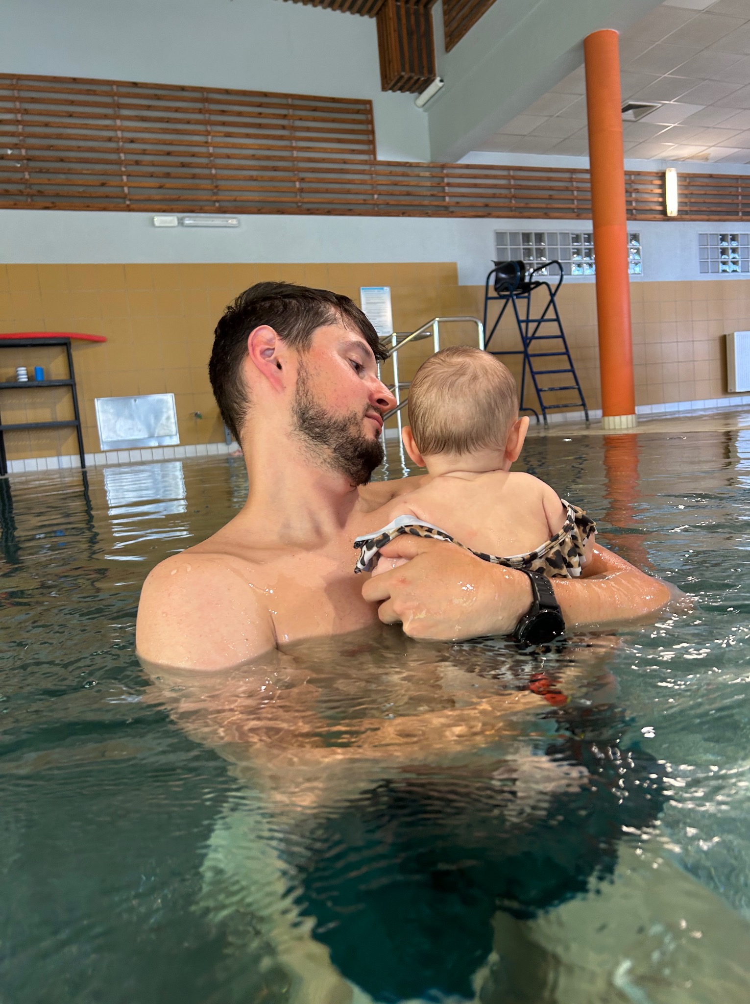 Mężczyzna z brodą trzymający małe dziecko w basenie, w tle drabinka ratownicza i drewniane panele ścienne.