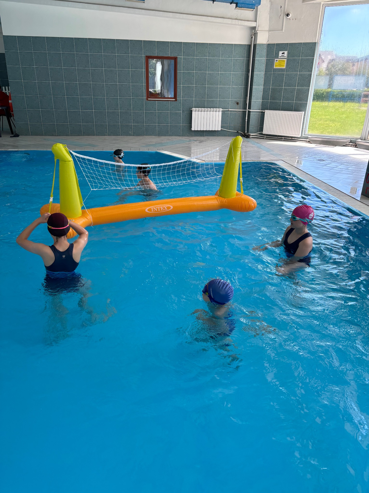 Lekcja pływania: grupa dzieci w czepkach i strojach kąpielowych w basenie z siatką do siatkówki wodnej. Zajęcia rekreacyjne w wodzie.