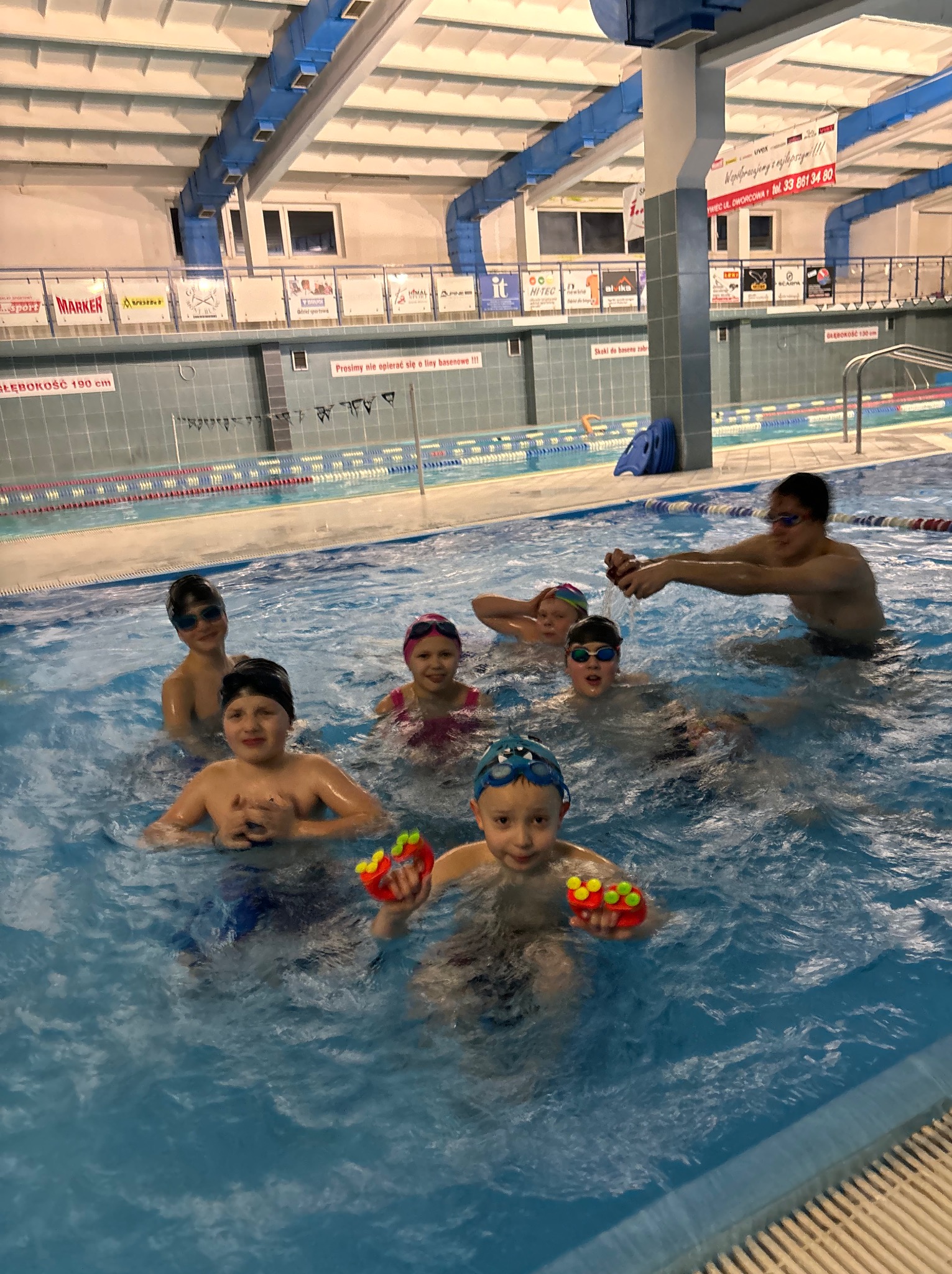 Grupa dzieci w basenie podczas zajęć. Chłopiec z przodu trzyma zabawki do wody. W tle widoczna ściana basenu z oznaczeniami głębokości.
