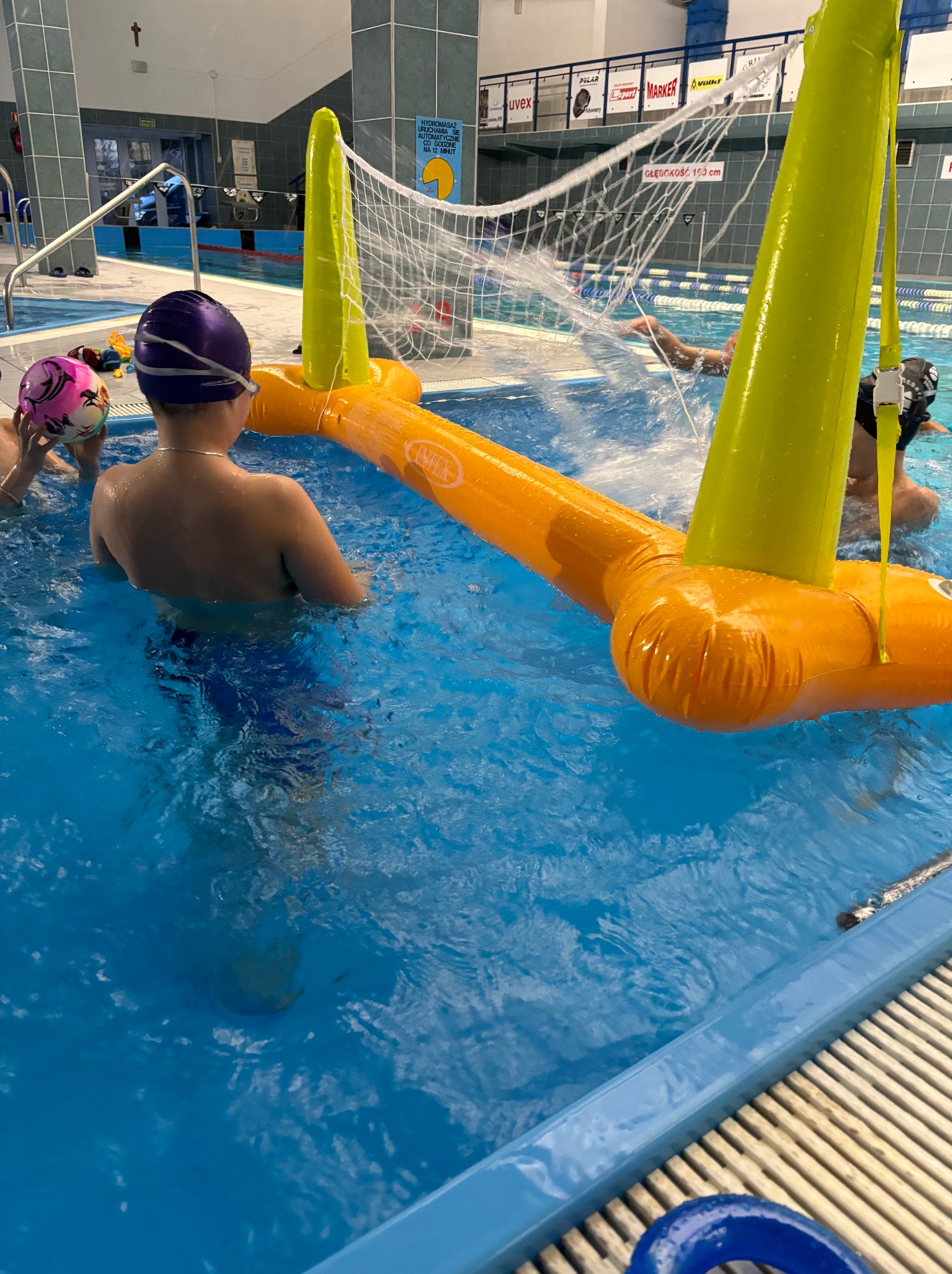 Dzieci w basenie podczas zajęć z piłki wodnej, z dmuchaną bramką. Chłopiec w czepku stoi tyłem, w tle osoba płynie pod wodą. Widoczna tablica informacyjna.