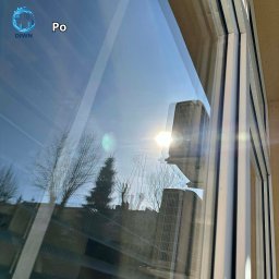 DIWN Nikodem Pańko - Błyszcząca szyba okienna po umyciu, odbijająca budynki i błękitne niebo. Widoczne ramy okienne i napis 'Po'.