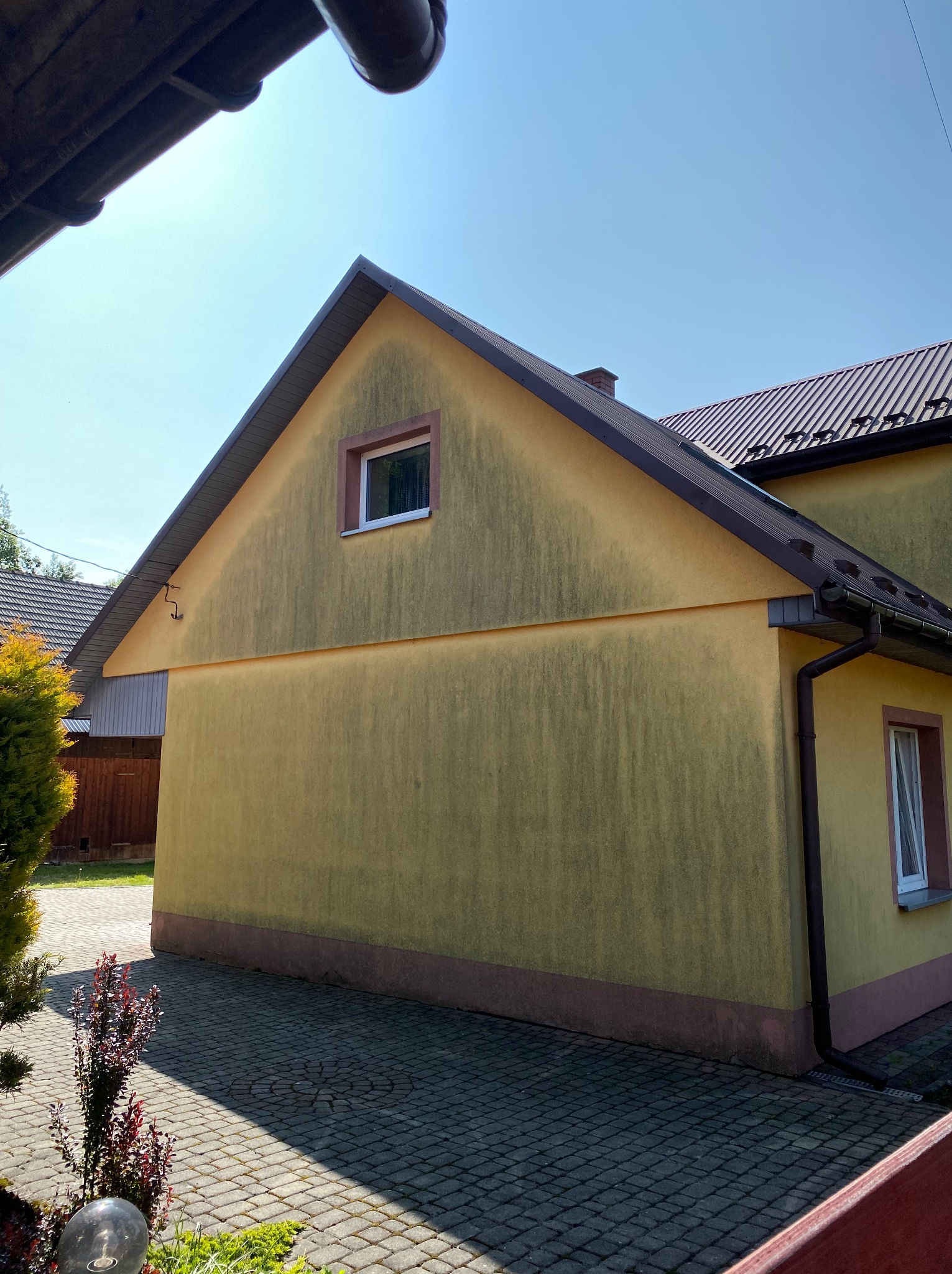 Żółta elewacja domu z widocznymi zabrudzeniami i zaciekami, ukazująca potrzebę mycia. Widoczny fragment dachu i okna.