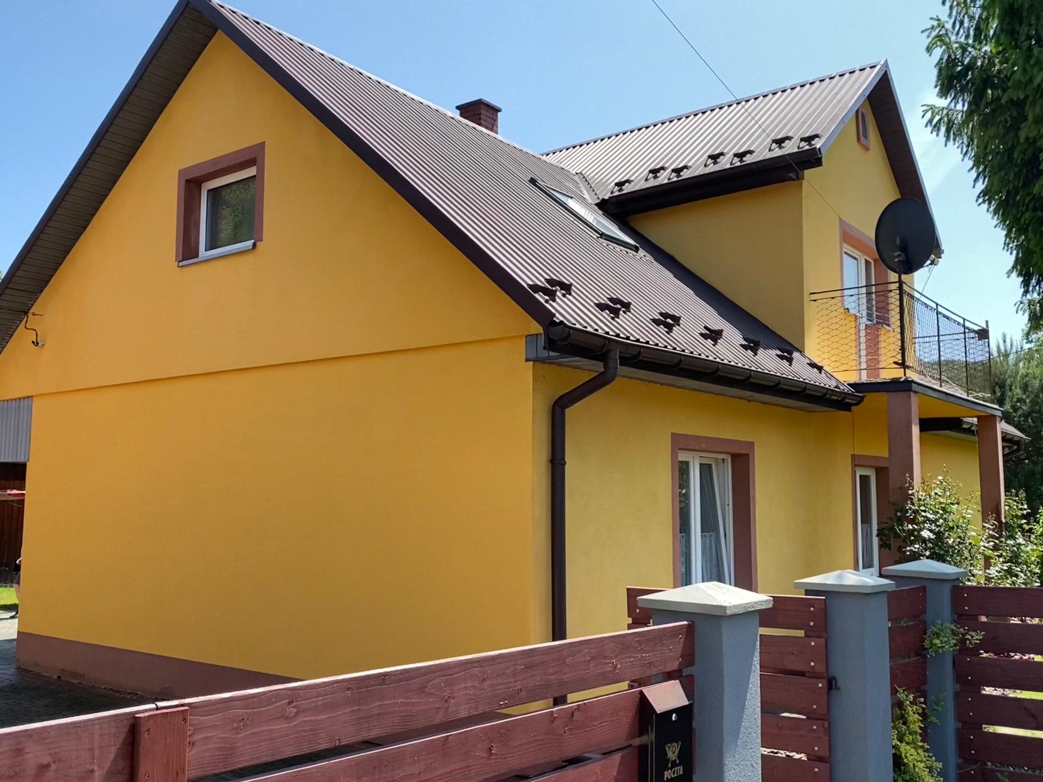 Żółty dom jednorodzinny z brązowym dachem i stolarką okienną, widoczny zza drewnianego ogrodzenia z skrzynką pocztową. Słoneczny dzień.