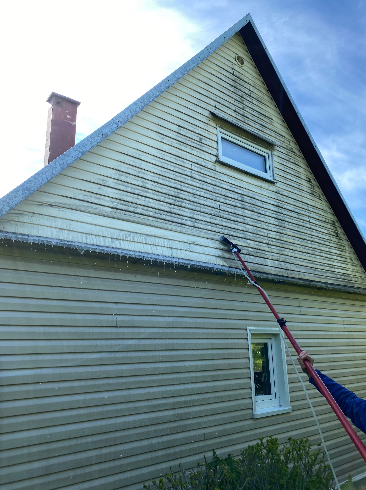 Mycie elewacji domu z sidingu za pomocą szczotki na teleskopowym kiju. Widoczne zacieki i spływająca woda. Częściowo umyta ściana budynku.