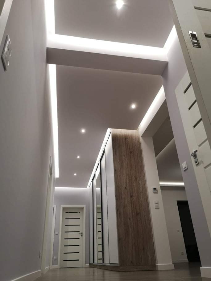 Nowoczesny przedpokój z oświetleniem LED w suficie podwieszanym i szafą wnękową z lustrami. Minimalistyczny design, szare ściany, białe drzwi.
