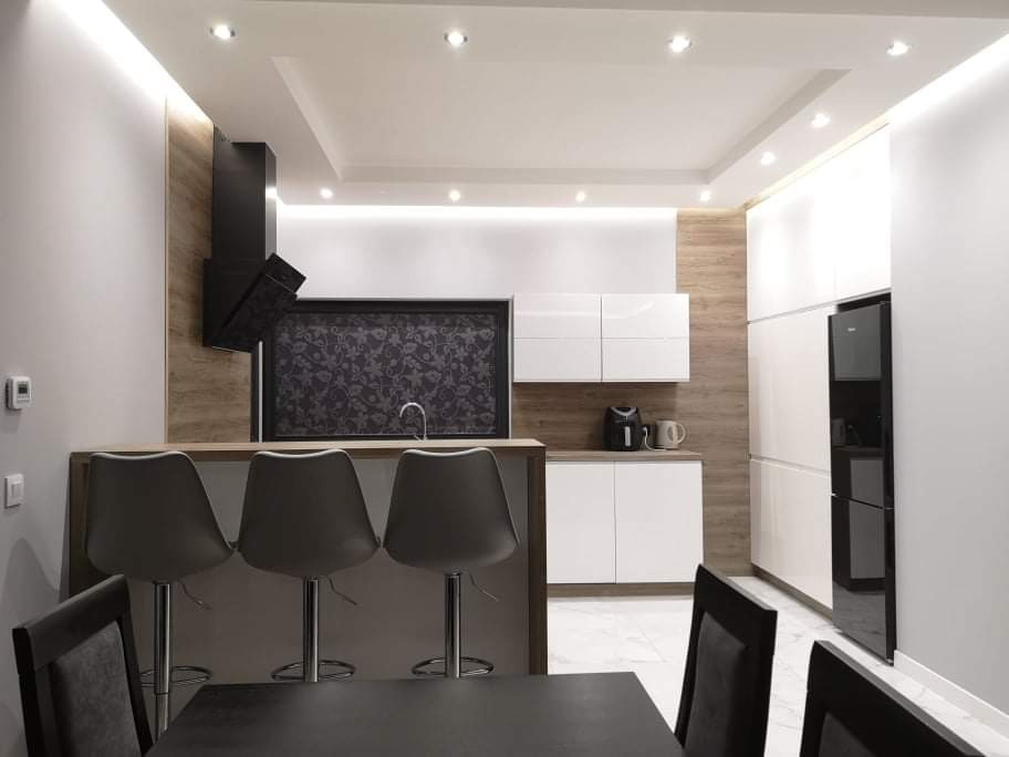 Nowoczesna kuchnia po remoncie: białe szafki, drewniane akcenty, czarny okap i wyspa z hokerami. Minimalistyczny design z oświetleniem LED.