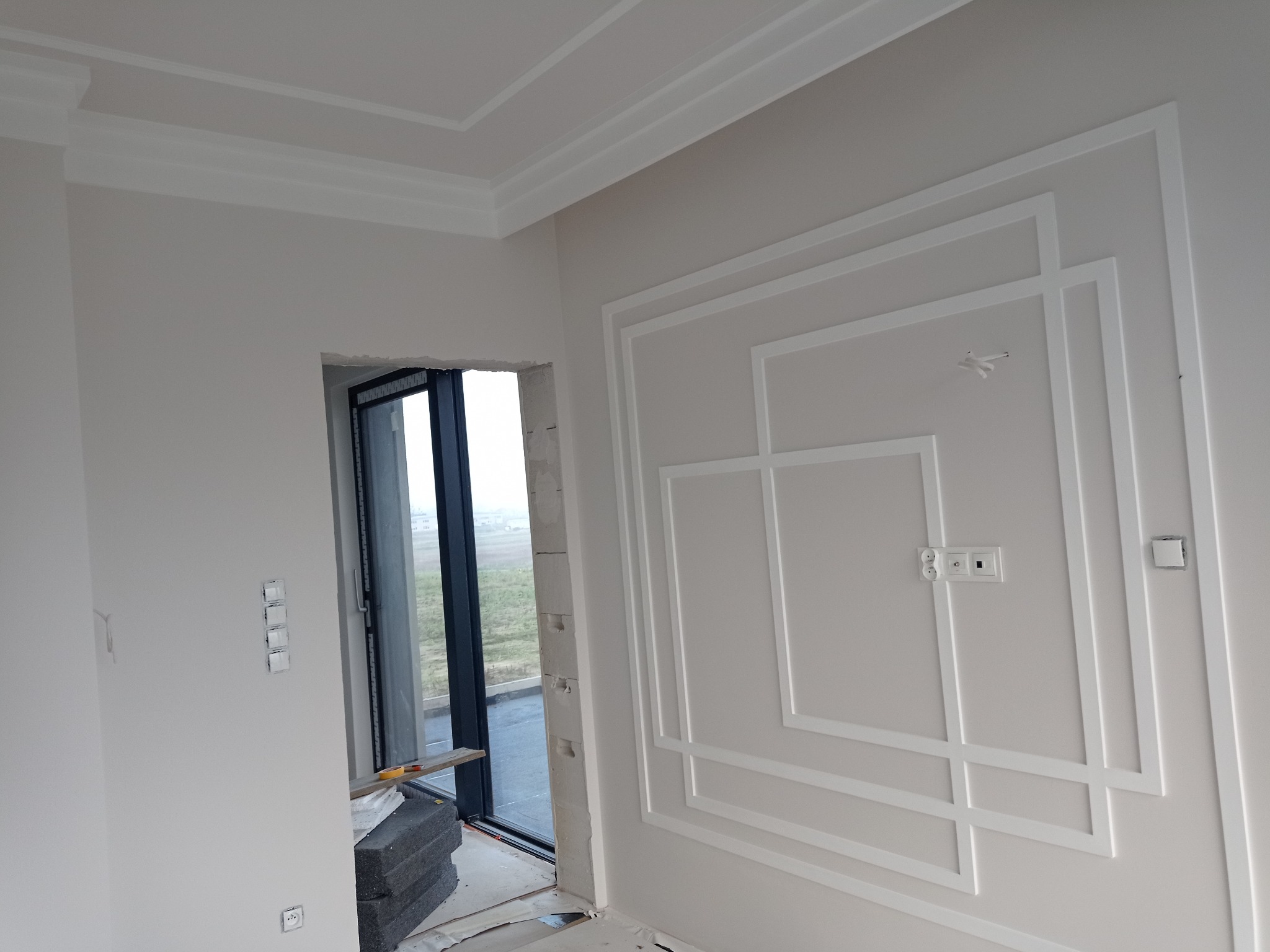 Białe listwy dekoracyjne na ścianie i suficie w jasnym wnętrzu, widok na okno i elementy instalacji elektrycznej. Klasyczny design w nowoczesnym wydaniu.