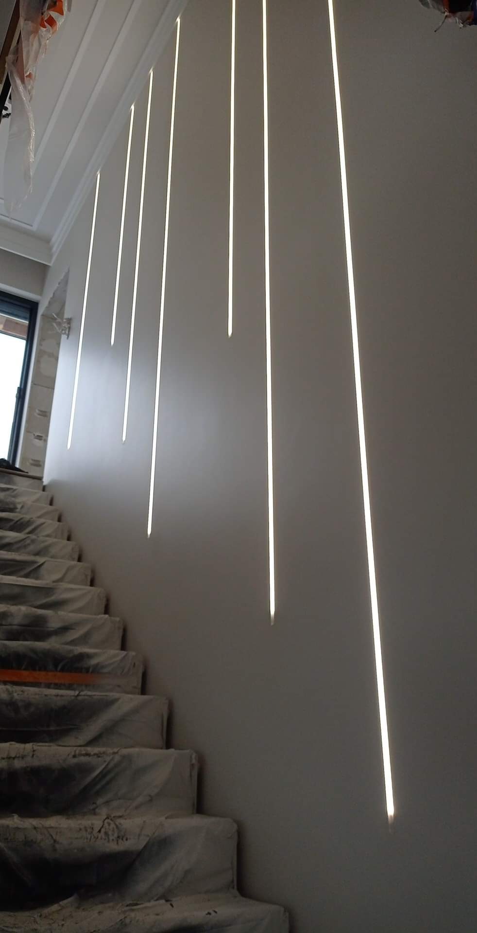 Nowoczesne oświetlenie schodów z pionowymi, wpuszczonymi w ścianę liniami LED. Schody zabezpieczone folią malarską. Minimalistyczny design, szare ściany, jasne światło.