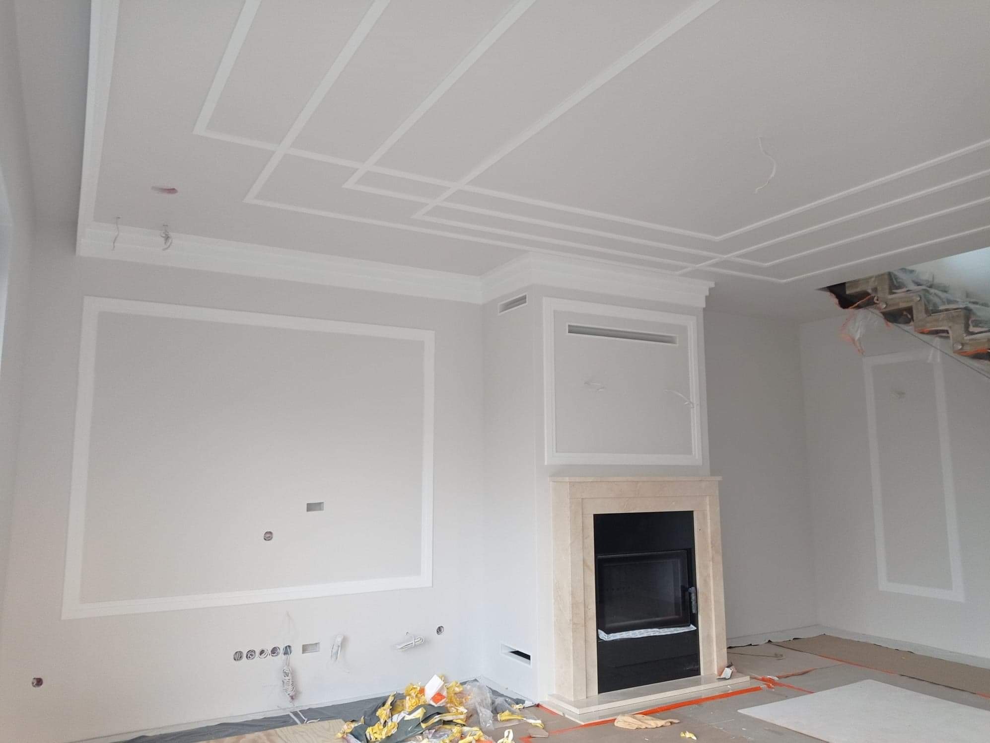 Jasne wnętrze z kominkiem i dekoracyjnymi listwami na ścianach i suficie. Widoczne ślady prac wykończeniowych, podłoga zabezpieczona folią.