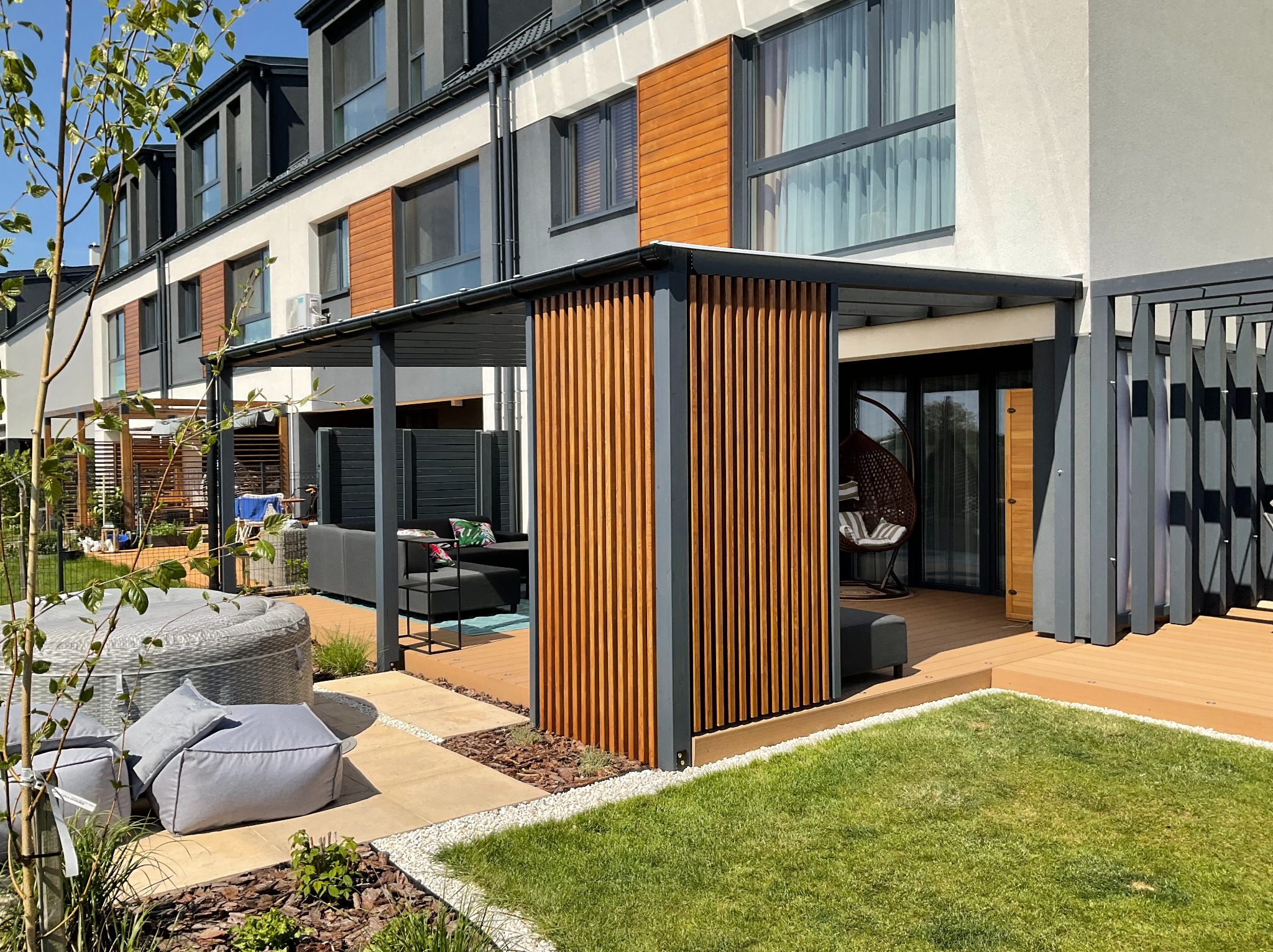 Nowoczesna pergola z drewnianymi lamelami i szarą konstrukcją na tarasie z kompozytu, z widokiem na meble ogrodowe i wiszący fotel. Słoneczny dzień.