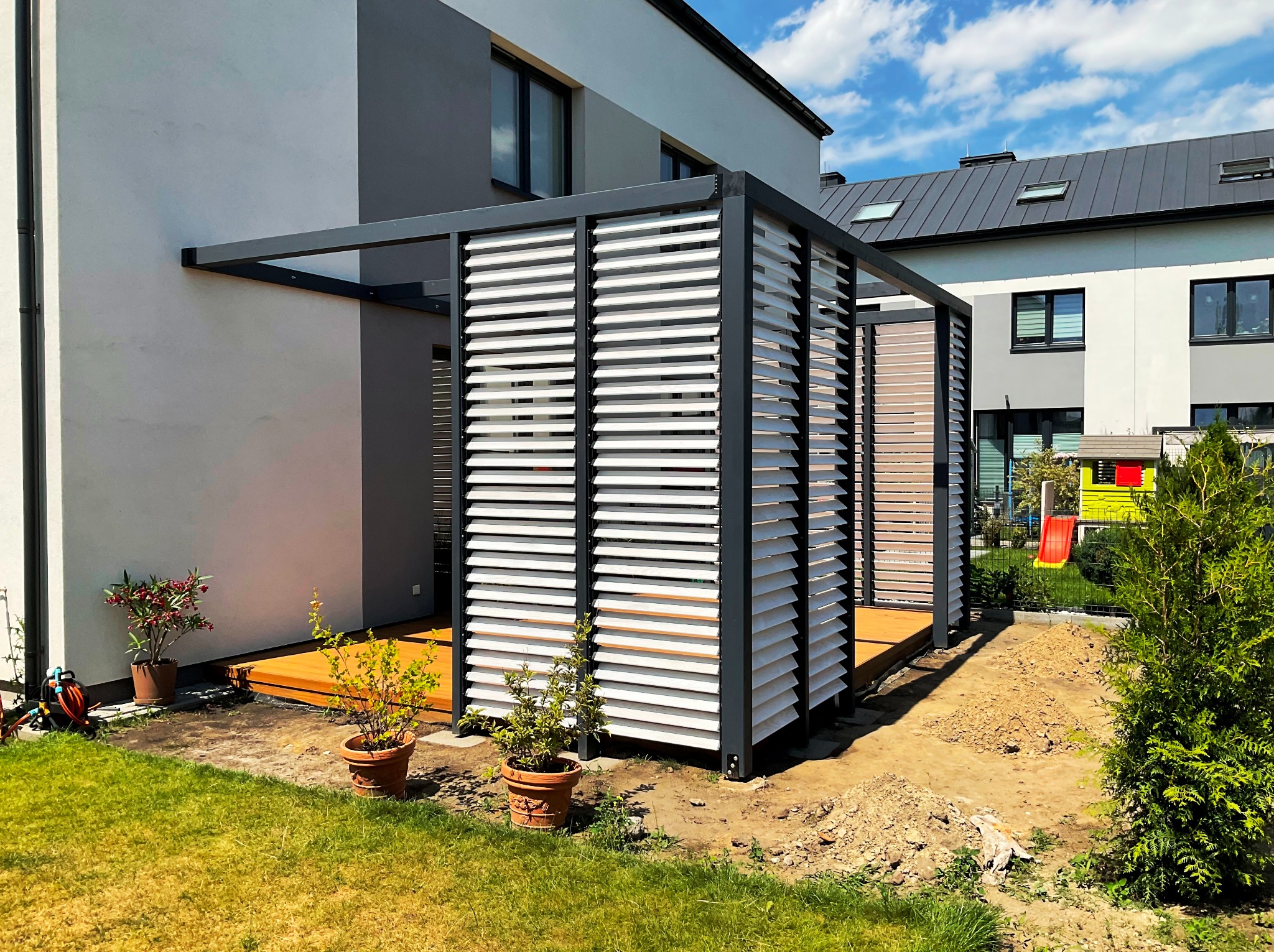 Nowoczesna pergola z szarymi ramami i białymi lamelami, przylegająca do budynku. Widoczny drewniany taras, rośliny w donicach i trawnik w słoneczny dzień.