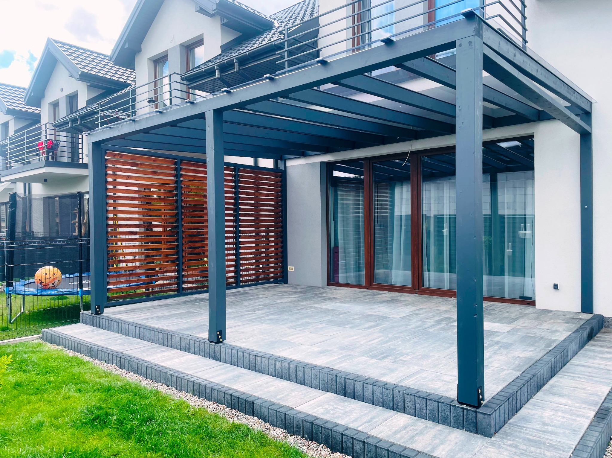 Nowoczesna, grafitowa pergola z drewnianą przesłoną na tarasie z szarej kostki. Widok na dom z balkonami i zielony trawnik. Elementy małej architektury.