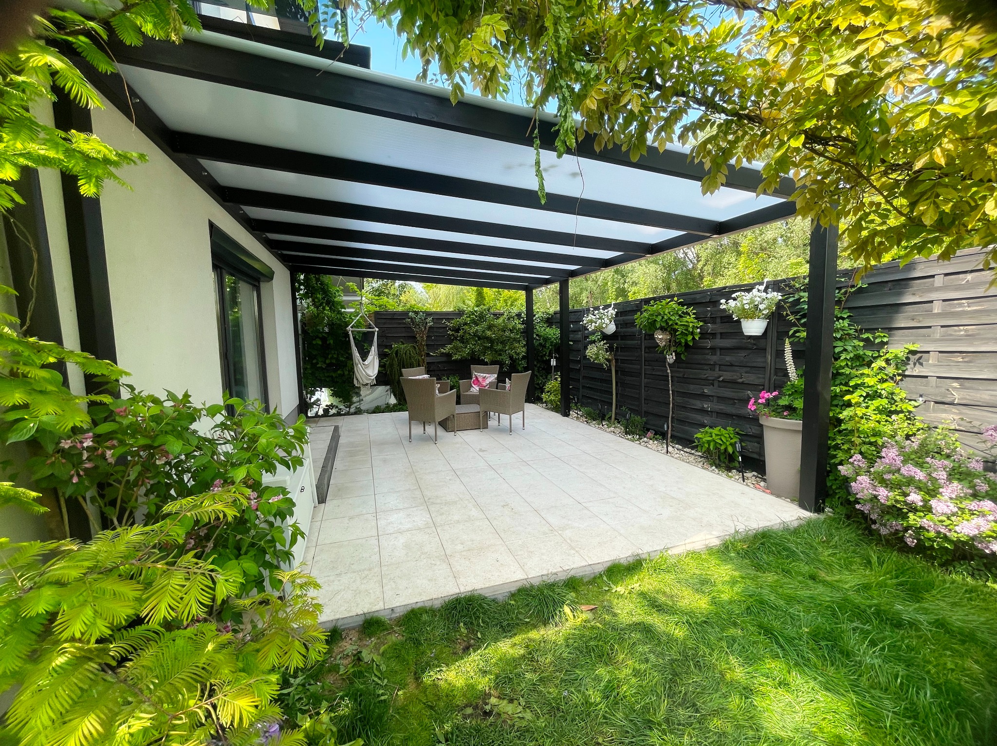 Nowoczesna pergola z czarną konstrukcją i jasnym zadaszeniem, osłaniająca taras z meblami ogrodowymi. Zieleń ogrodu i dekoracje kwiatowe tworzą przytulną przestrzeń.