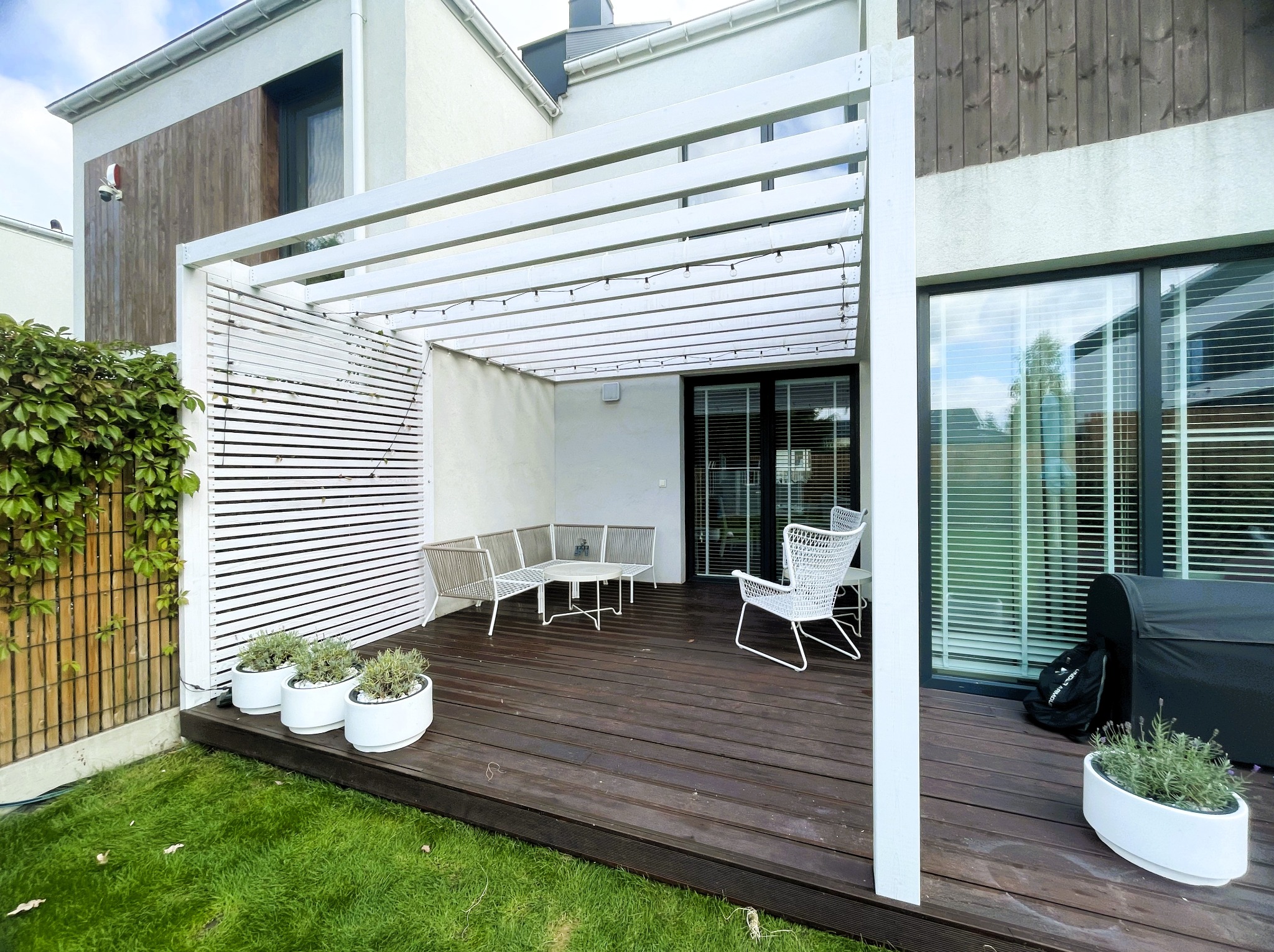 Nowoczesna, biała konstrukcja z lameli nad tarasem z ciemnego drewna. Minimalistyczne meble ogrodowe i donice z roślinami. Tło stanowi jasny budynek.