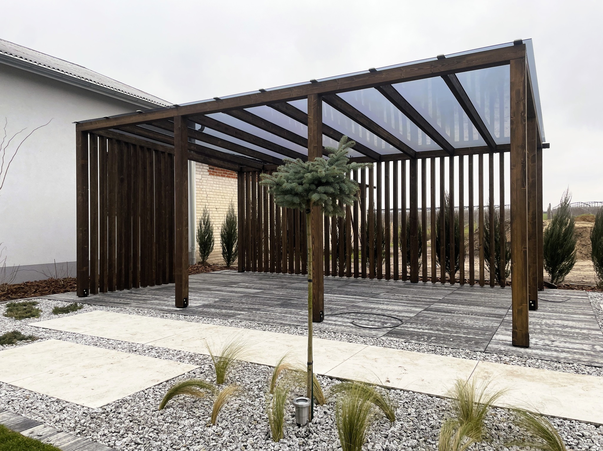 Nowoczesna, ciemna pergola z przezroczystym zadaszeniem na tarasie z szarych płyt. W tle widoczny budynek z białej cegły i zielone rośliny.