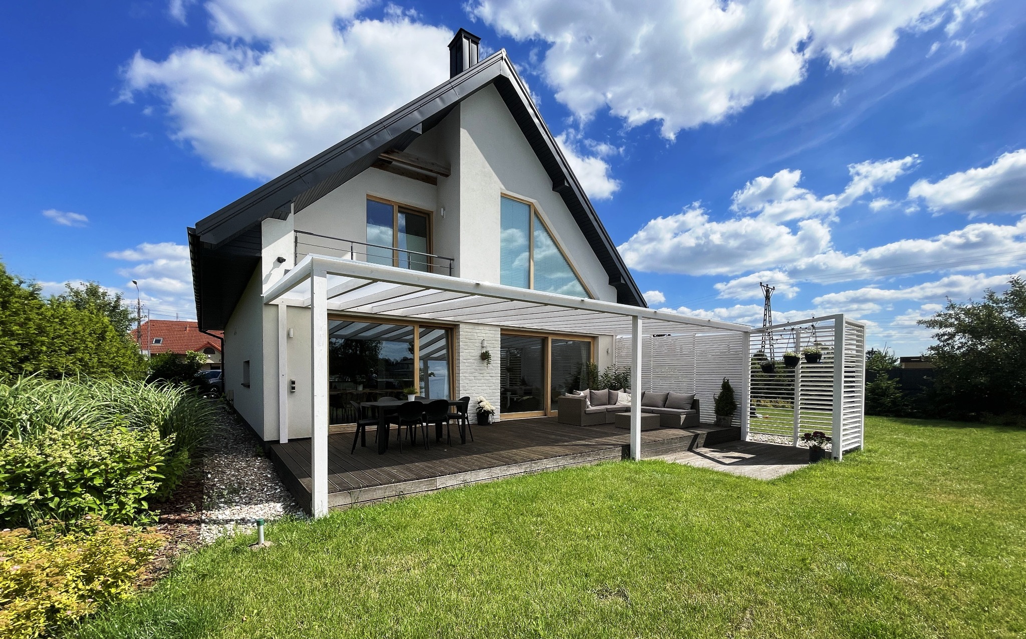 Nowoczesny taras z drewnianą podłogą i białą pergolą przy domu. Meble ogrodowe, dekoracyjne panele. Błękitne niebo z chmurami w tle. Słoneczny dzień.