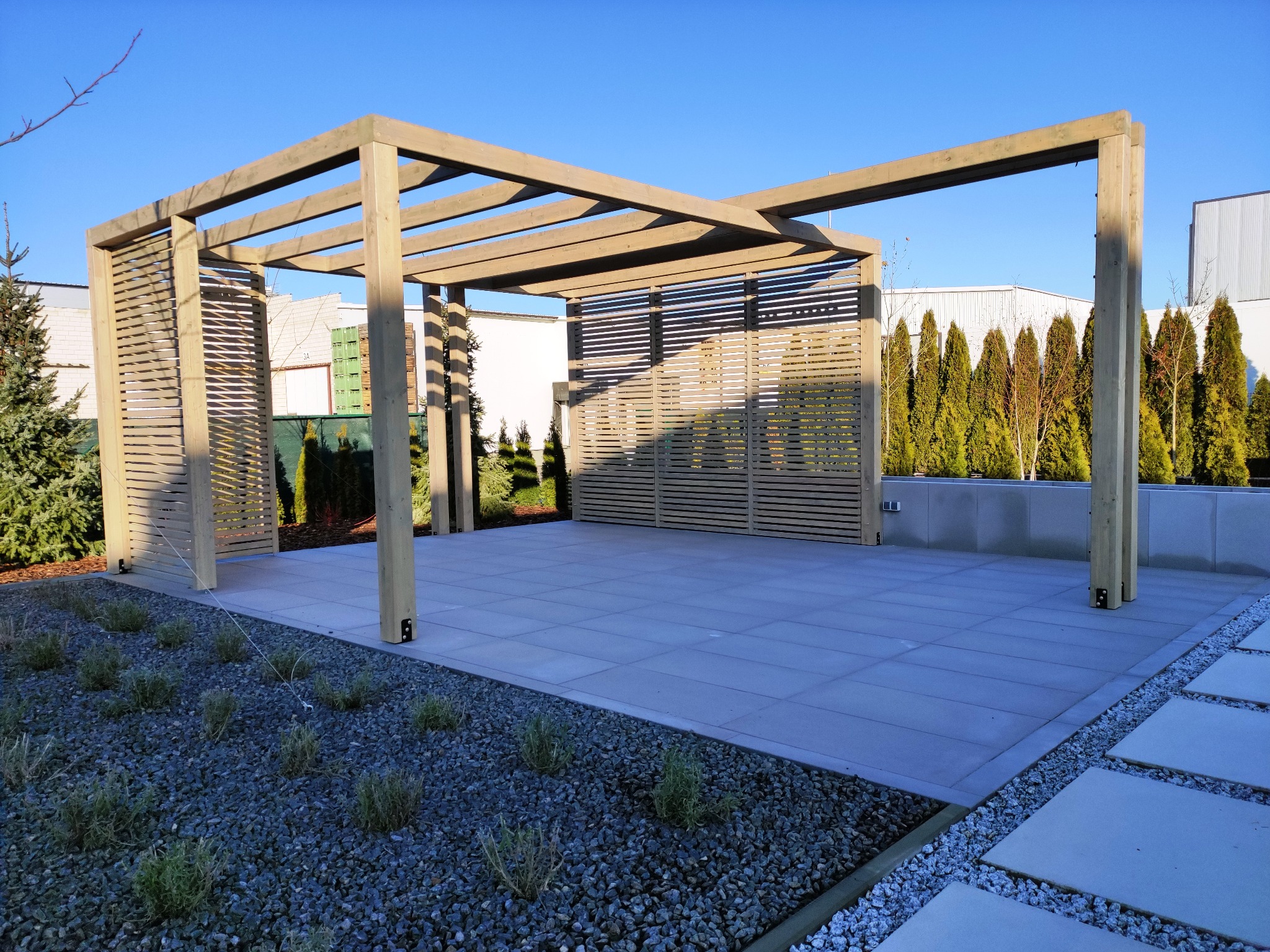 Nowoczesna pergola z drewnianą konstrukcją i ażurowymi ścianami, otoczona szarym brukiem i zielenią. Minimalistyczny design w słoneczny dzień.