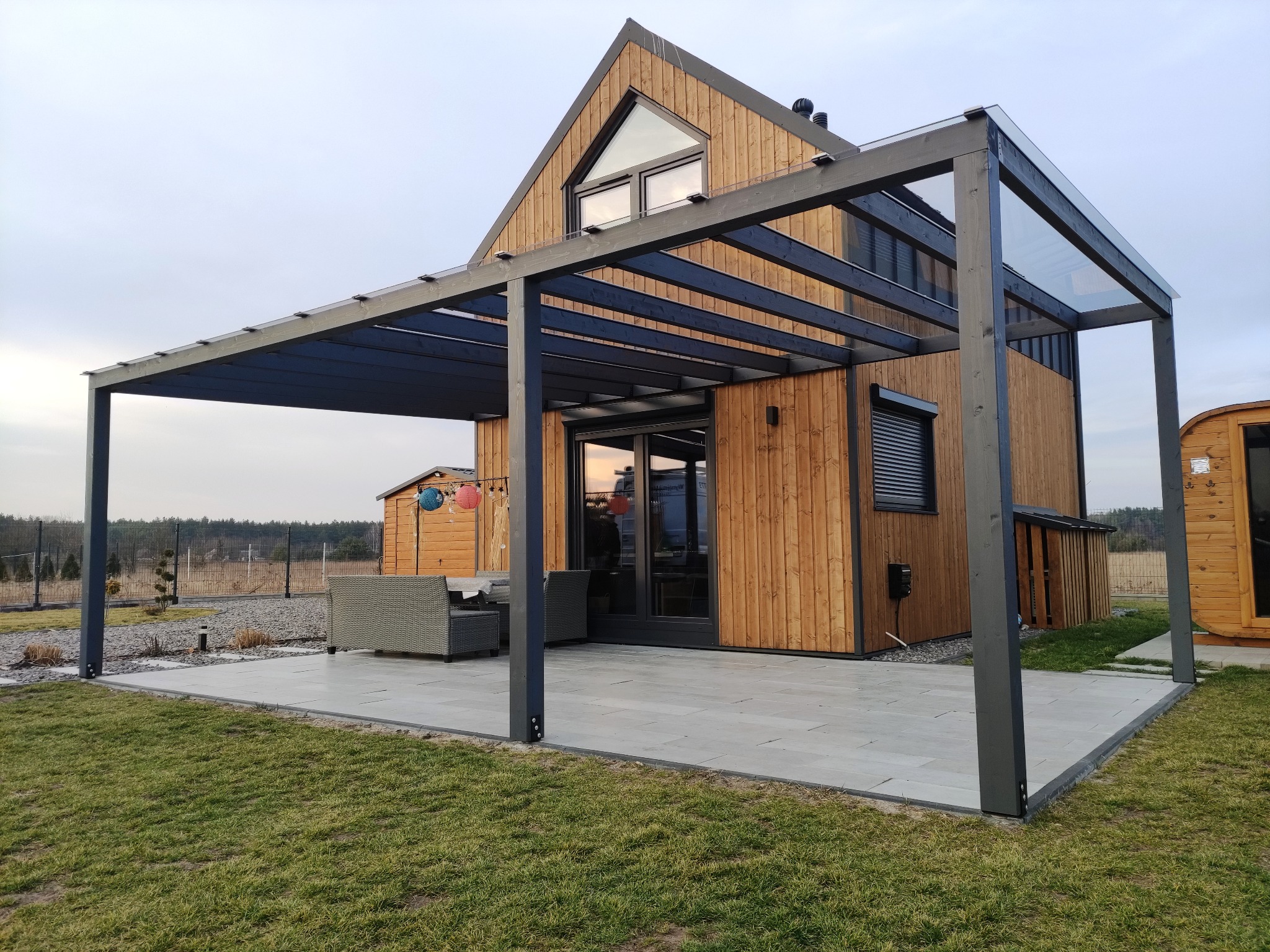 Nowoczesna, grafitowa pergola z przeszklonym dachem, przylegająca do drewnianego domu z dużymi oknami. Na tarasie zestaw mebli wypoczynkowych.